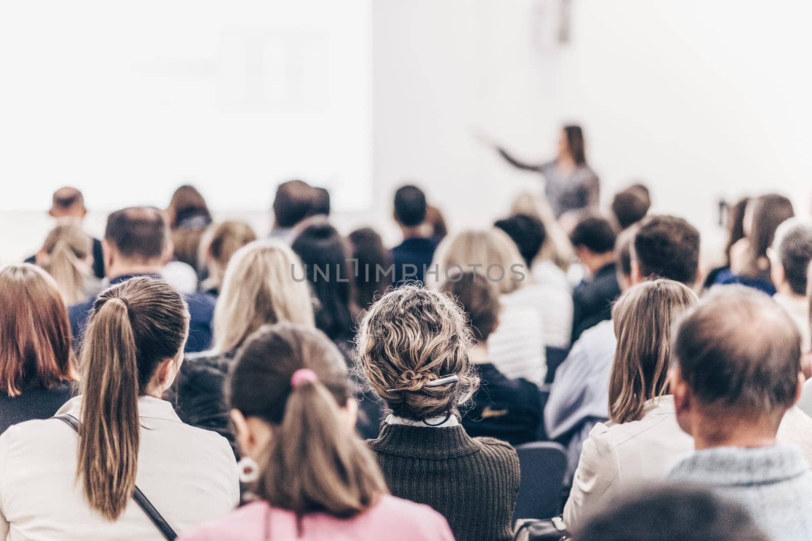 Business and entrepreneurship symposium. Female speaker giving a talk at business meeting. Audience in conference hall. Rear view of unrecognized participant in audience.