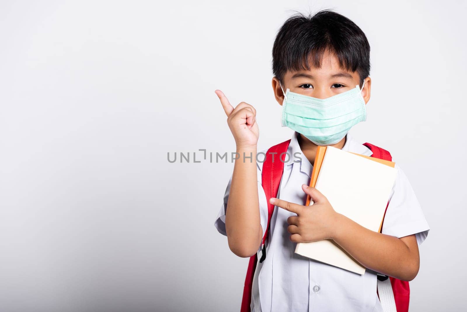 Asian student kid boy wear student thai uniform and protect face mask ready to go to school pointing finger to space in studio shot isolated on white background, preschool, new normal back to school