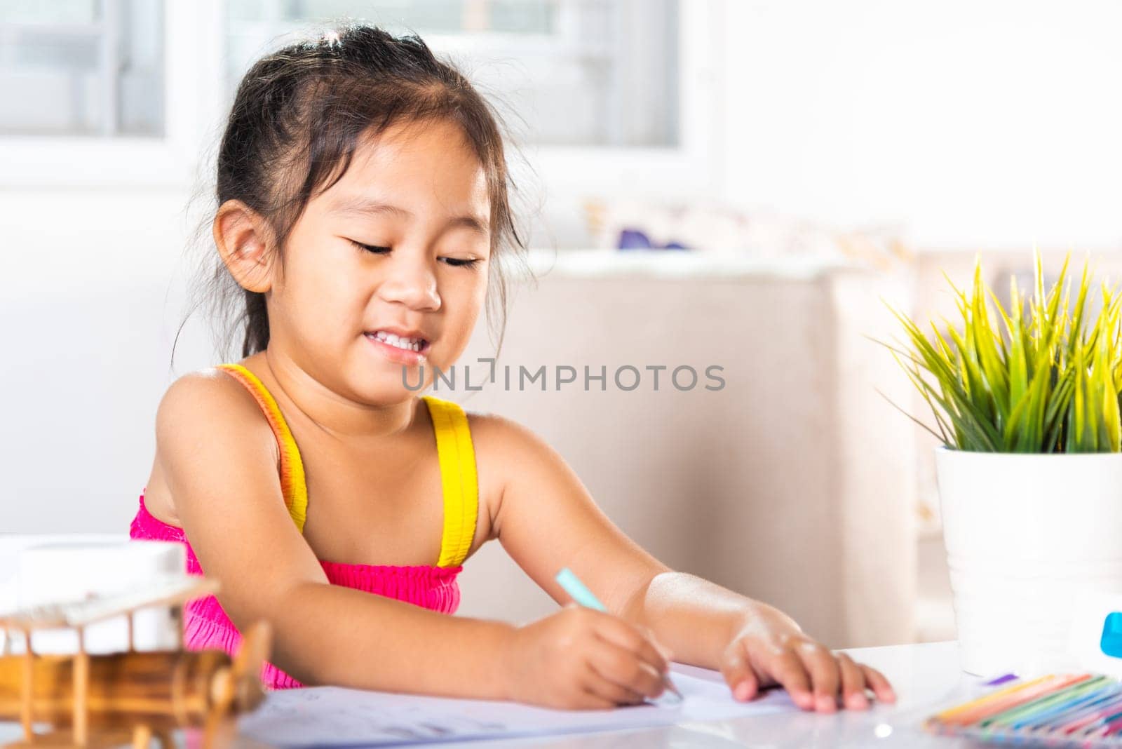 Asian cute kid preschooler sitting on table smiling she draw and writing picture with pencil at home to learning arts homework, Happy child little girl drawing cartoon on paper before paint the color
