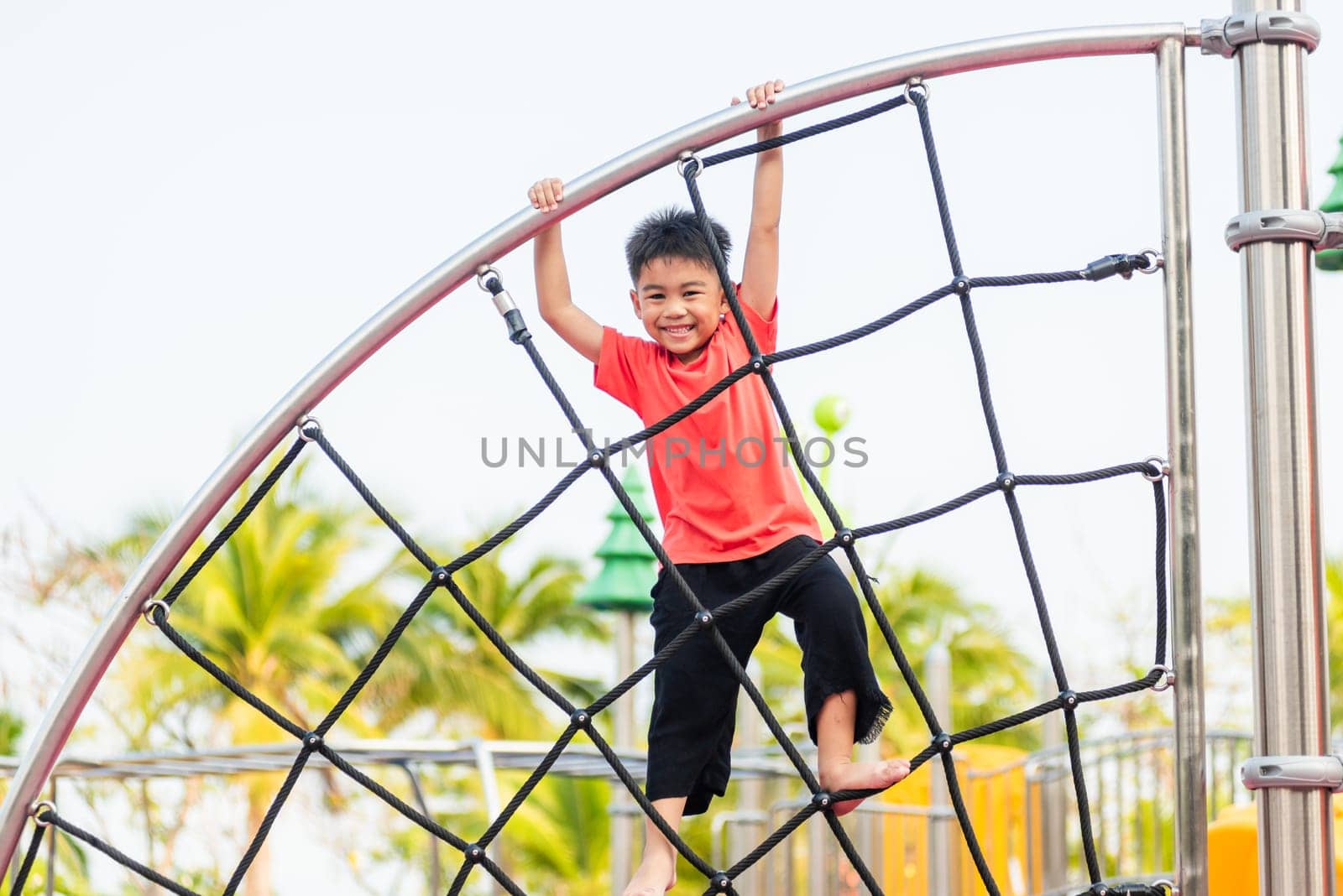 Asian child smiling playing climbing outdoor playground, happy preschool little kid having funny while playing climbs on a rope climbing net on playground equipment in daytime in summer, Little boy