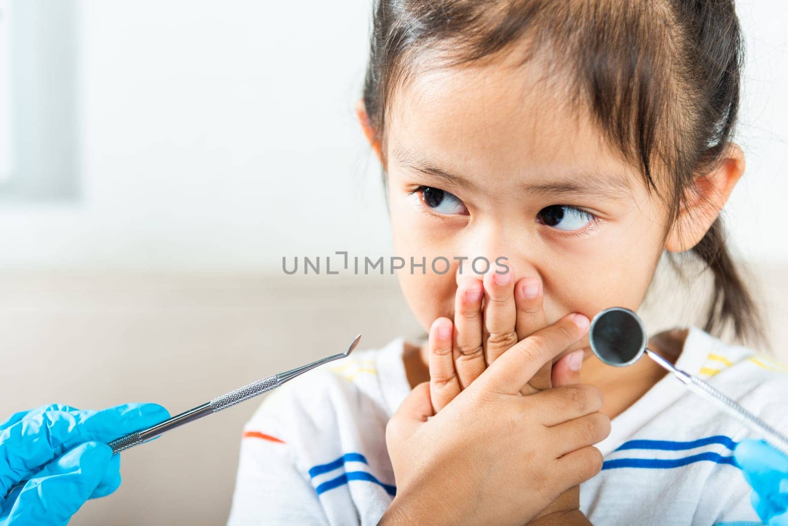Dental kid examination. Doctor examines oral cavity of child uses mouth mirror to check teeth cavity but little girl cover mouth no need to checking she afraid, scared and closes his mouth with hands
