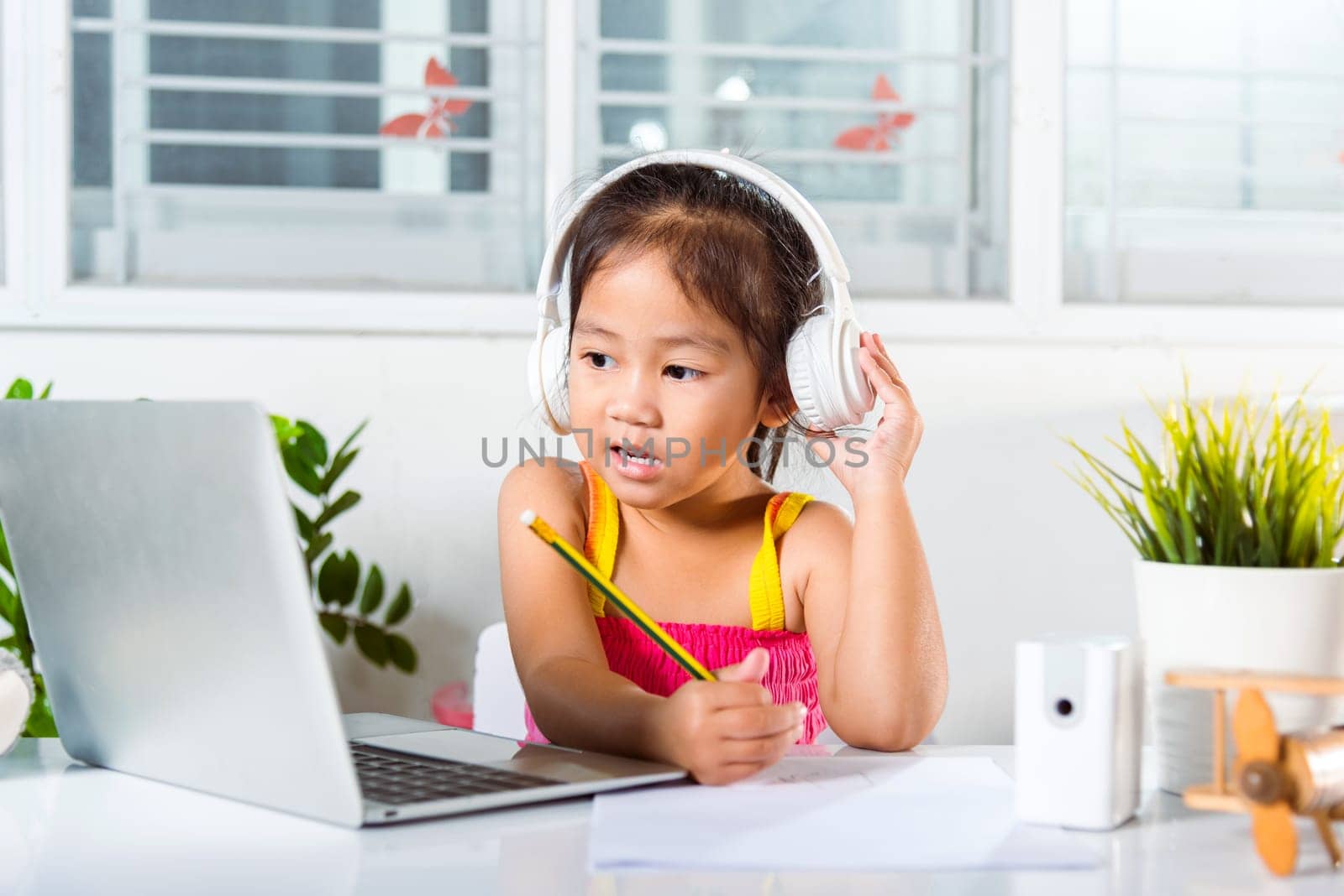 Asian child girl studying video conference distant education at home. Little kid preschool wear headphones sit at desk use laptop computer and communicates on Internet online video call with teacher