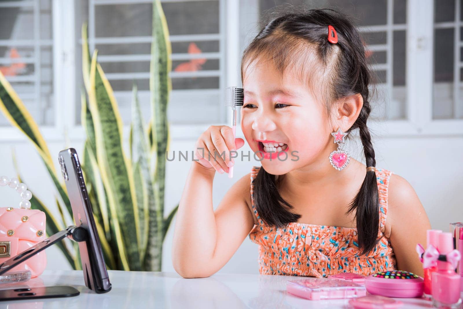 Asian funny little girl making makeup her face she look in smartphone and use eyebrow mascara, Learning activity to be woman, social media blogger recording vlog with cosmetics, online tutorial
