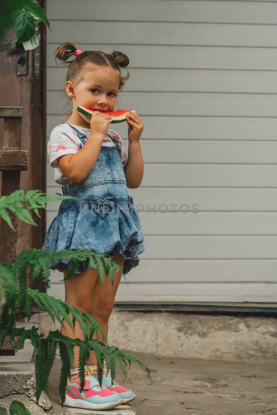 Concentrated little girl 3-4 years old with two ponytails eats watermelon. Child outdoors against background of building holds sweet healthy fruit in her hands. Summer, harvest, vitamins