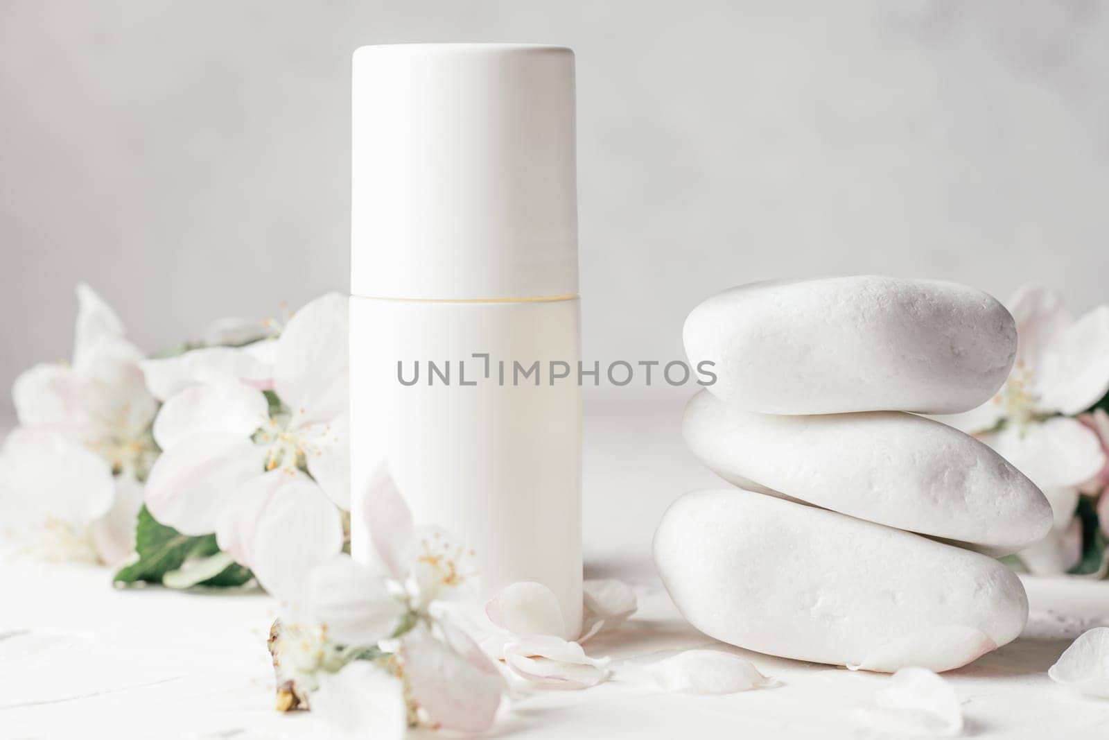Antiperspirant roll-on deodorant near stack of white pebble stones on light plaster surface with apple flowers