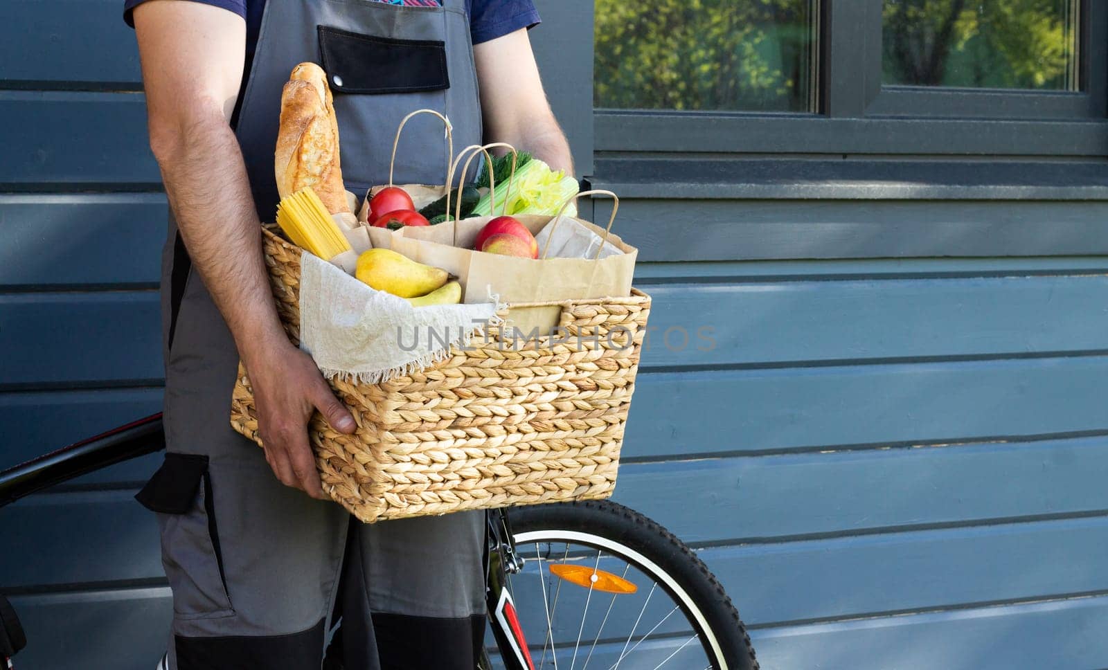 Fresh organic bread, vegetables, greens and fruits, cereals and pasta in a wicker basket in the hands of a bicycle courier. Bike eco-friendly delivery or donation of ecological farm food concept.