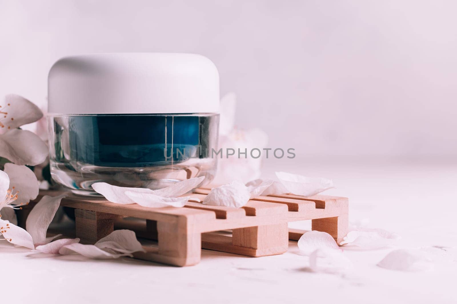Blue glass jar of cream on wooden podium in form of pallet on light plaster surface with apple flowers. Copy space