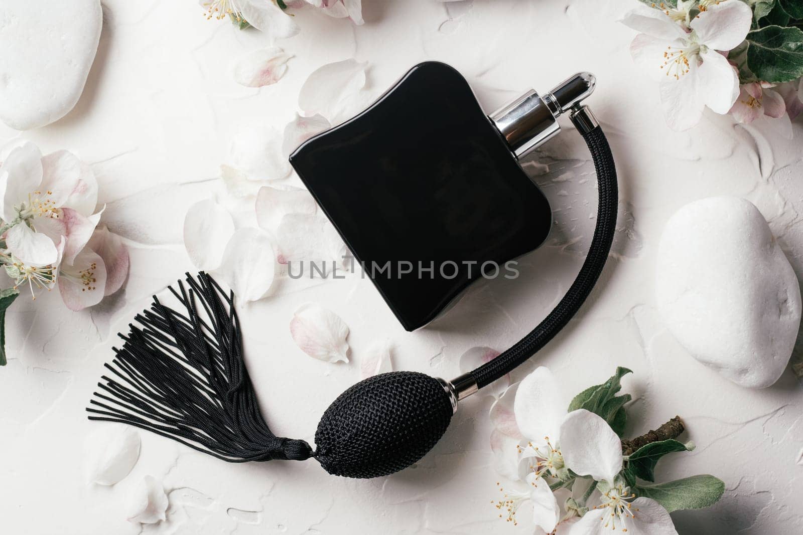 Black bottle eau de toilette or perfume with long tassel spray pomp on delicate background of apple flowers, petals and pebble stones lying on white gypsum plaster. Top view