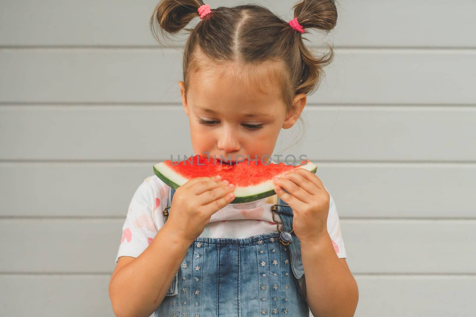 Concentrated little girl 3-4 years old with two ponytails eats watermelon. Child outdoors against background of building holds sweet healthy fruit in her hands. Summer, harvest, vitamins