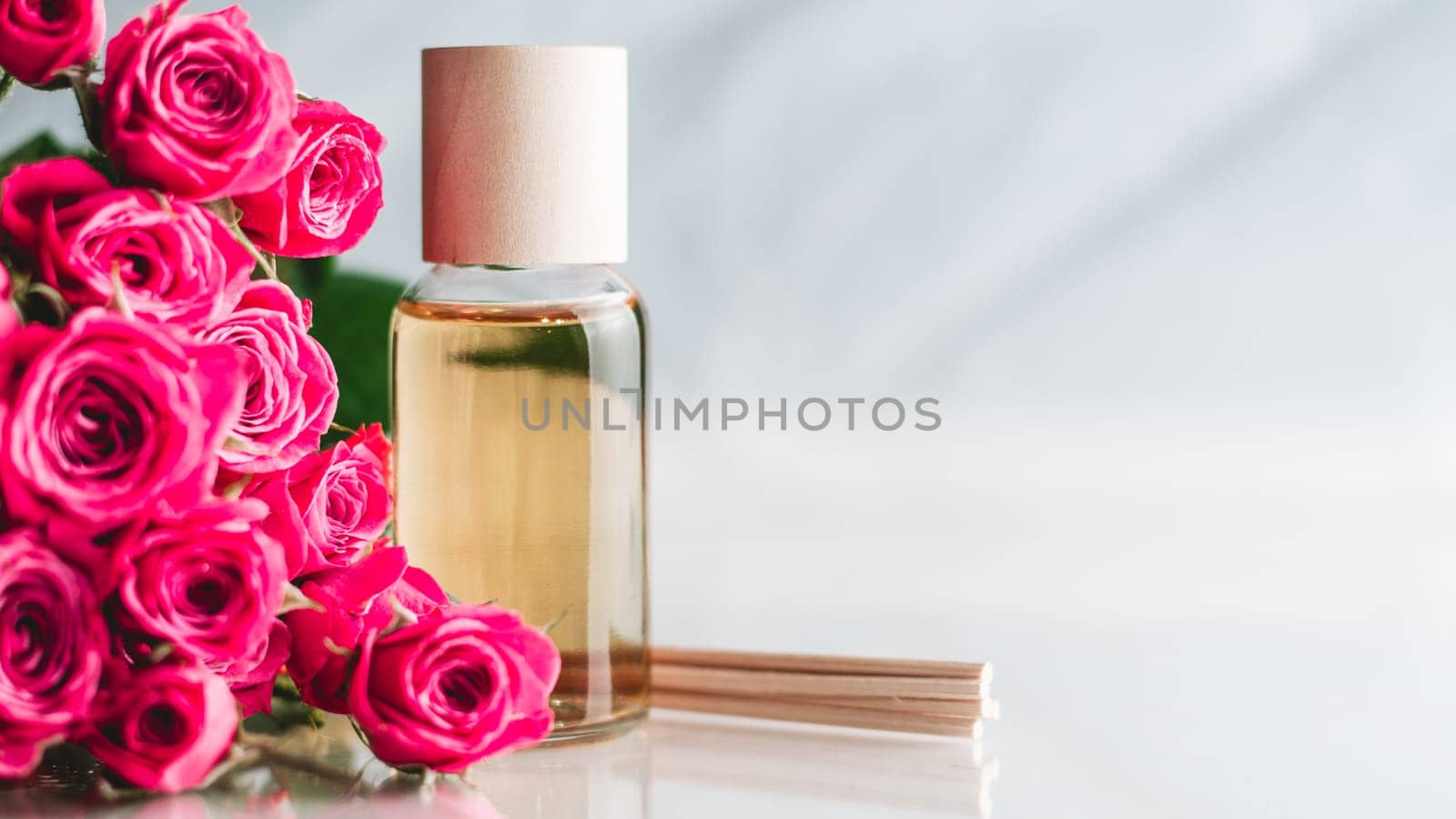 Clear bottle of yellow oil or perfume with wooden sticks and delicate pink roses on white surface. SPA salon and procedures concept. Banner. Copy space