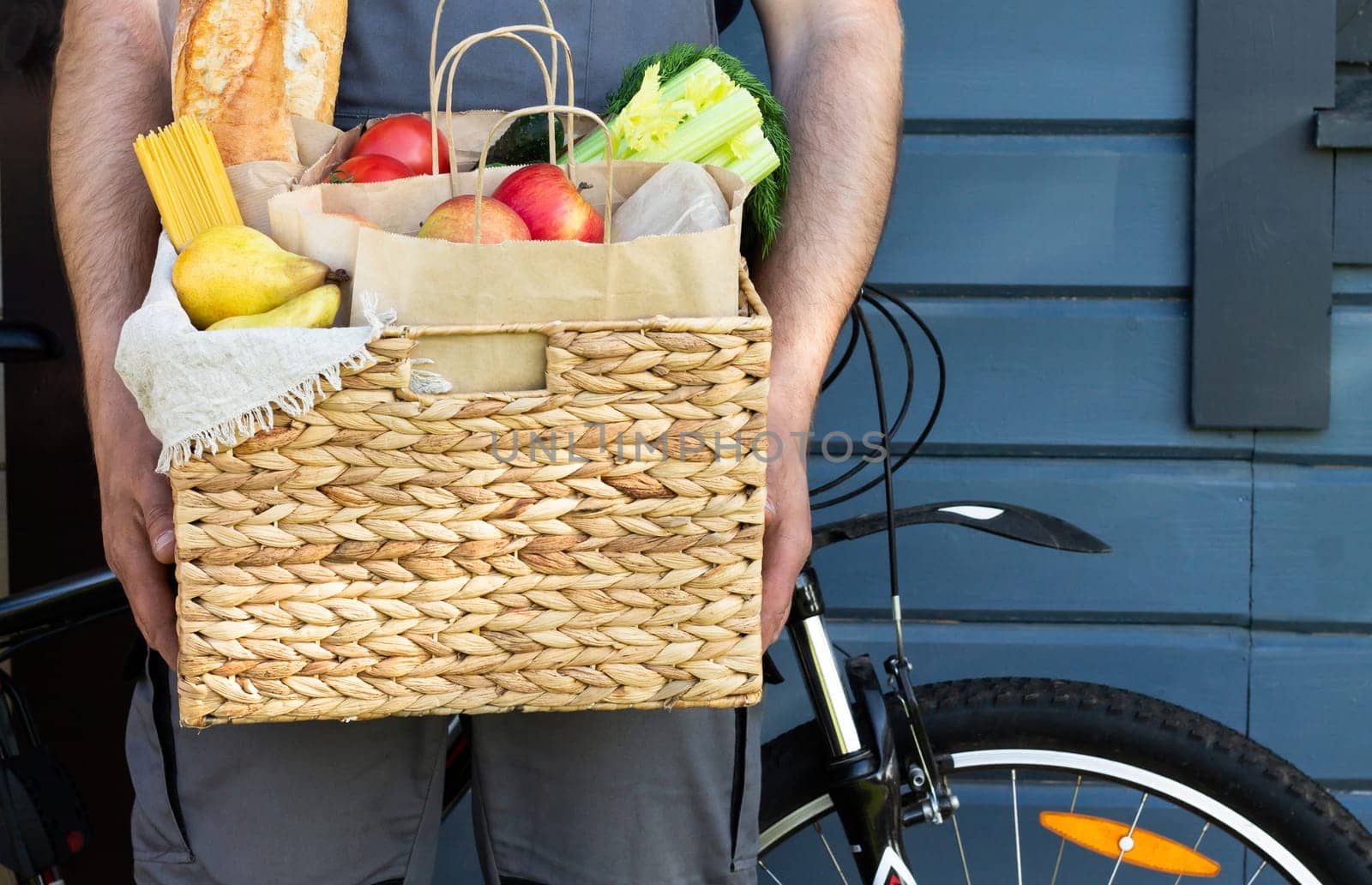 Fresh organic bread, vegetables, greens and fruits, cereals and pasta in a wicker basket in the hands of a bicycle courier. Bike eco-friendly delivery or donation of ecological farm food concept.