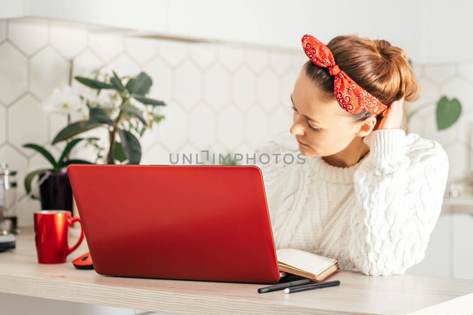Young woman sits with red laptop at home in kitchen in red bandage and white sweater and looks at monitor. Home work or study concept