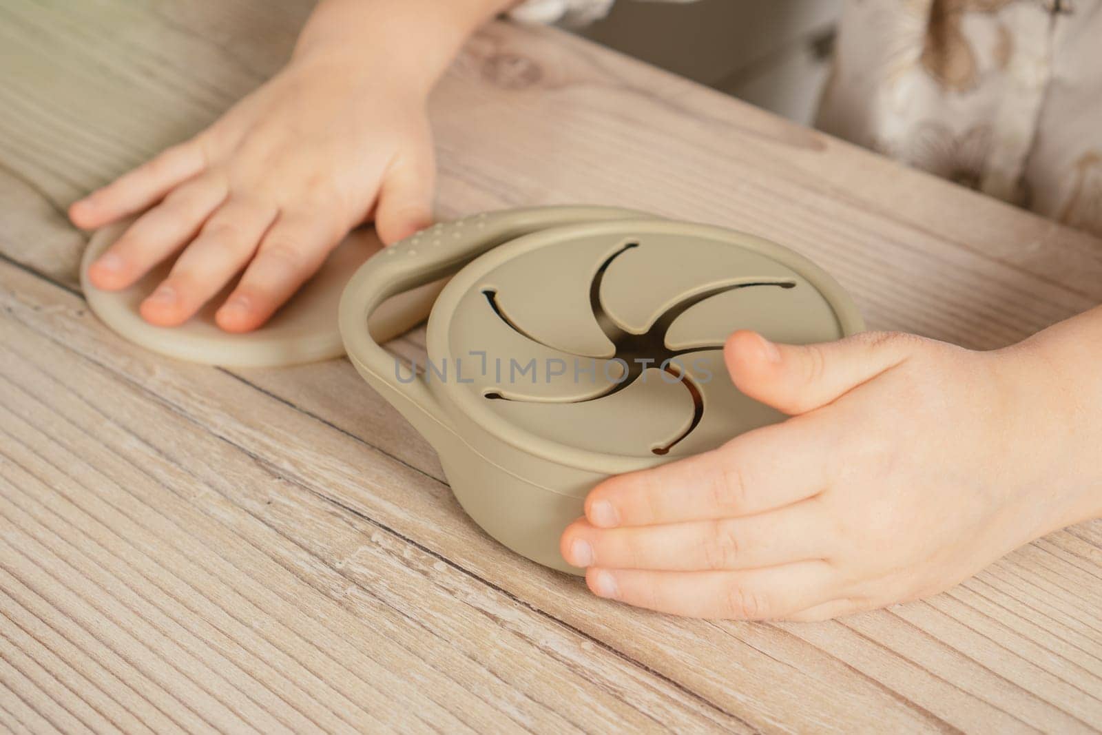 Unrecognizable child holding pastel gray silicone snack cup near lid at wooden table. Baby accessories, tableware, first feeding concept