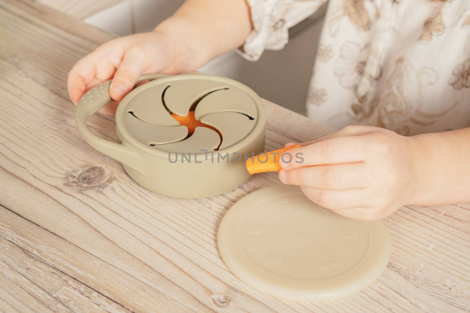 Unrecognizable child taking piece of carrot out from pastel gray silicone snack cup near lid at wooden table. Baby accessories, tableware, first feeding concept