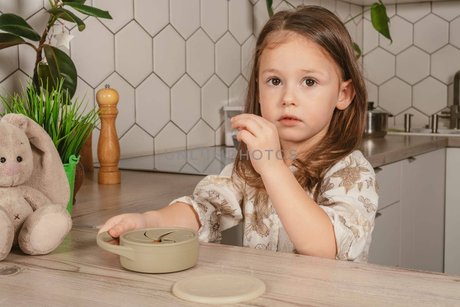 Little dark haired girl 3-4 years old taking piece of food out from pastel gray silicone snack cup and looking into frame in kitchen. Toy bunny nearby. Baby accessories, tableware, feeding concept