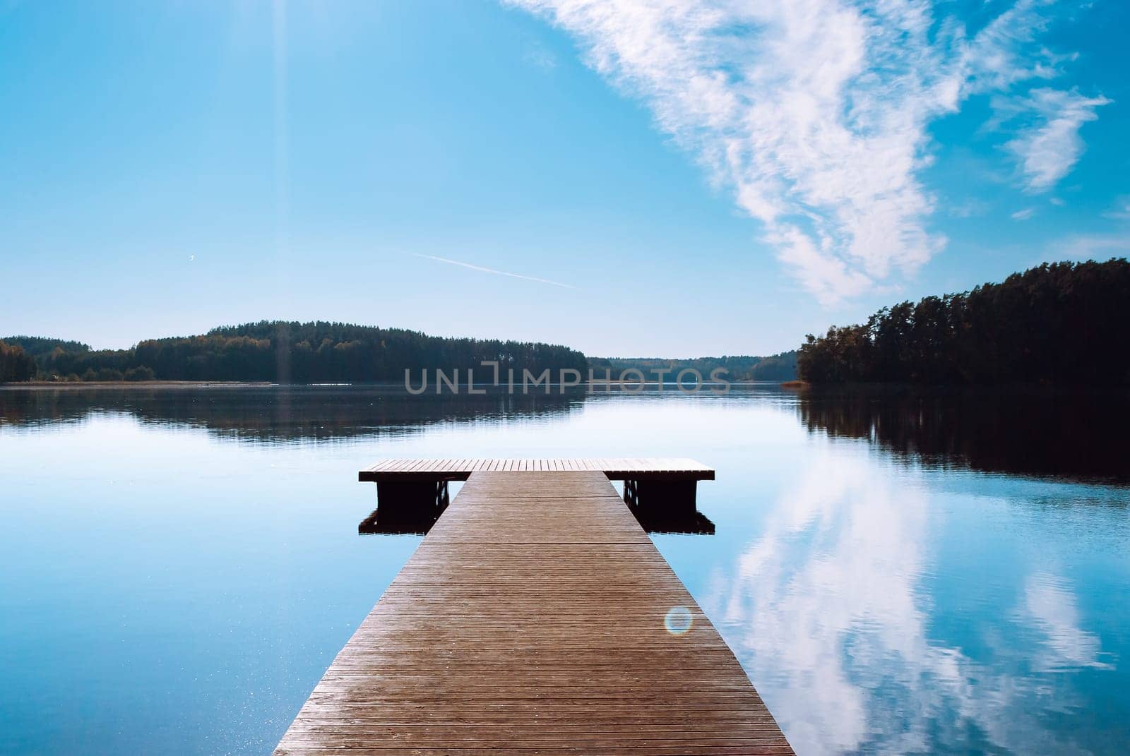 A wooden pier against the backdrop of a lake in the middle of the forest. Blue sky with clouds reflected in the water of Lake Baltieji Lakajai in Labanoras Regional Park.