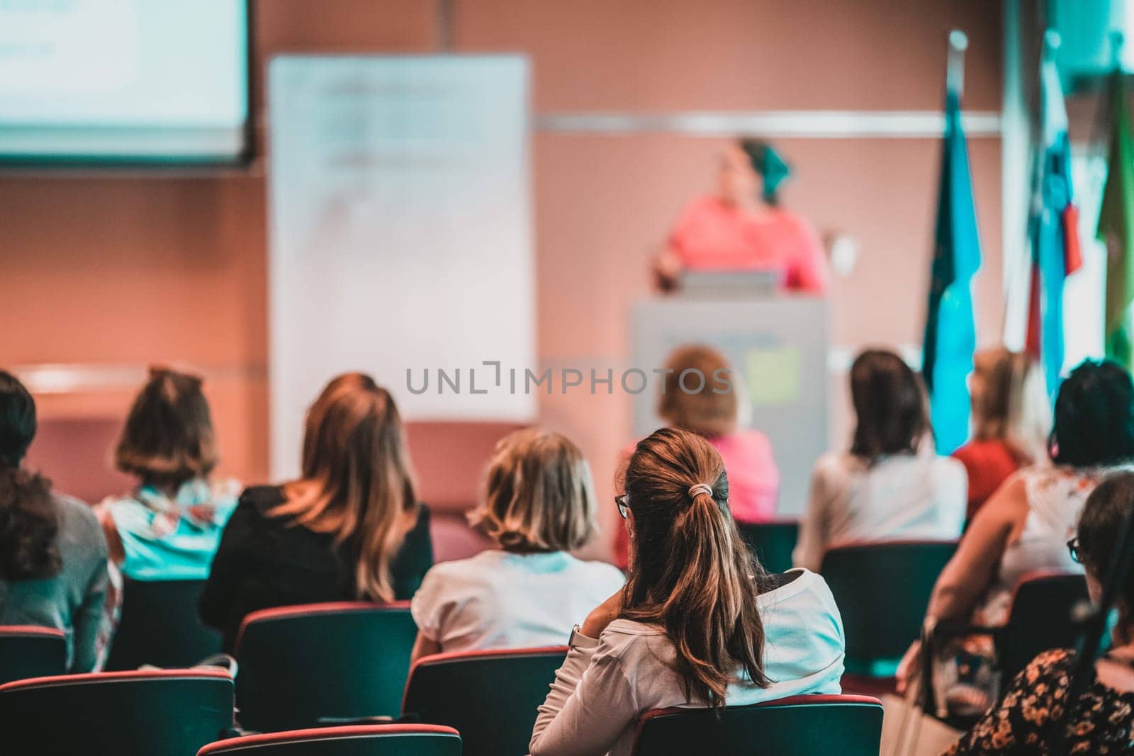 Business and entrepreneurship symposium. Female speaker giving a talk at business meeting. Audience in conference hall. Rear view of unrecognized participant in audience.