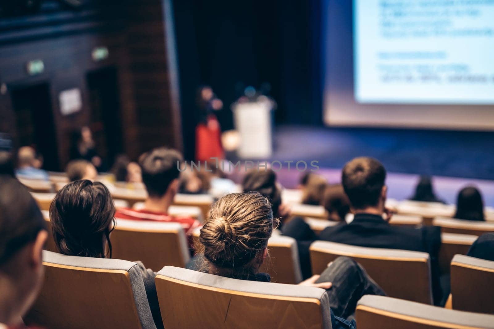 Business and entrepreneurship symposium. Female speaker giving a talk at business meeting. Audience in conference hall. Rear view of unrecognized participant in audience.