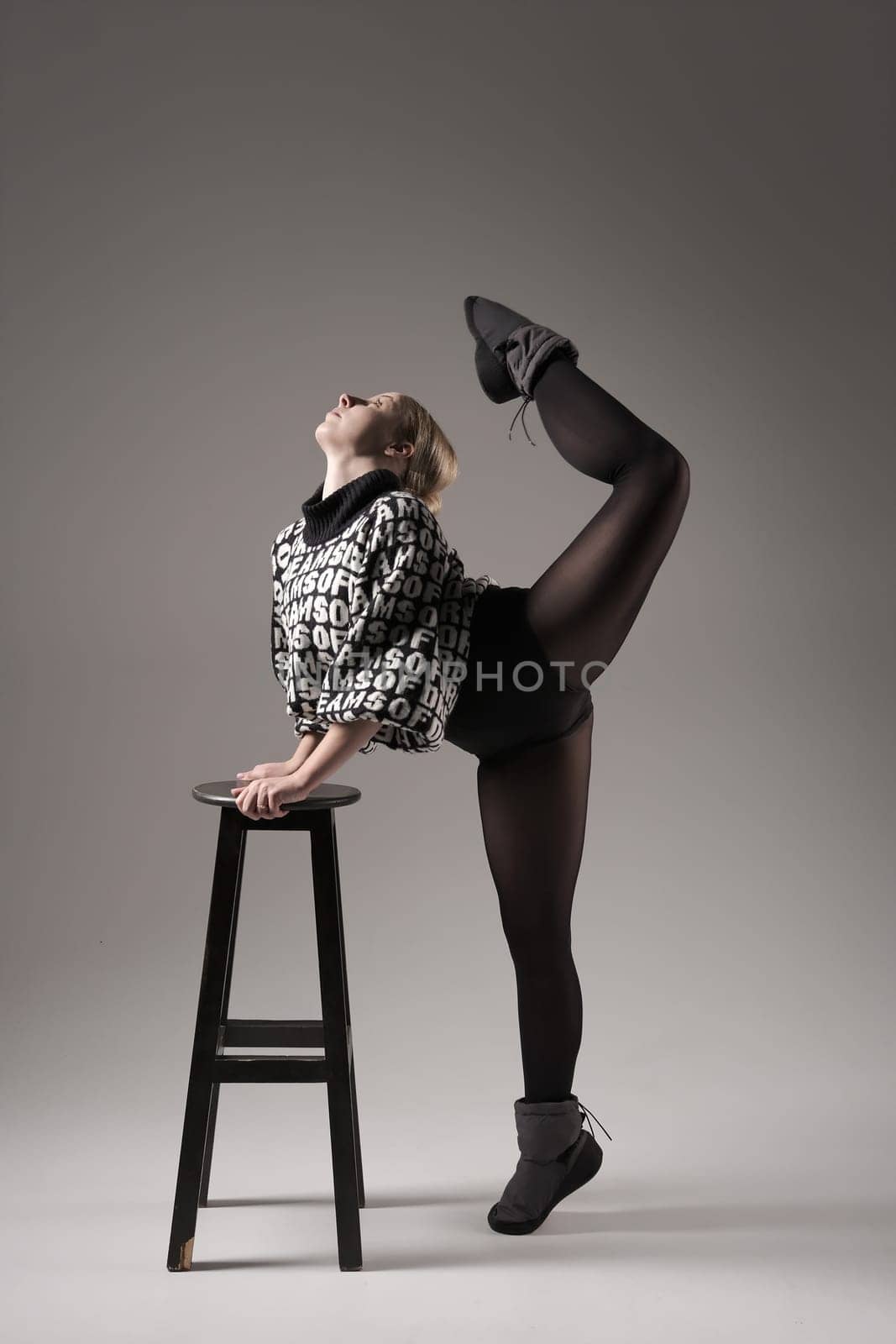 Ballet dancer woman black dress on gray background, Ballerina posing and showing her flexibility on gray background in studio with chair. Text on sweater DREAMS OF.