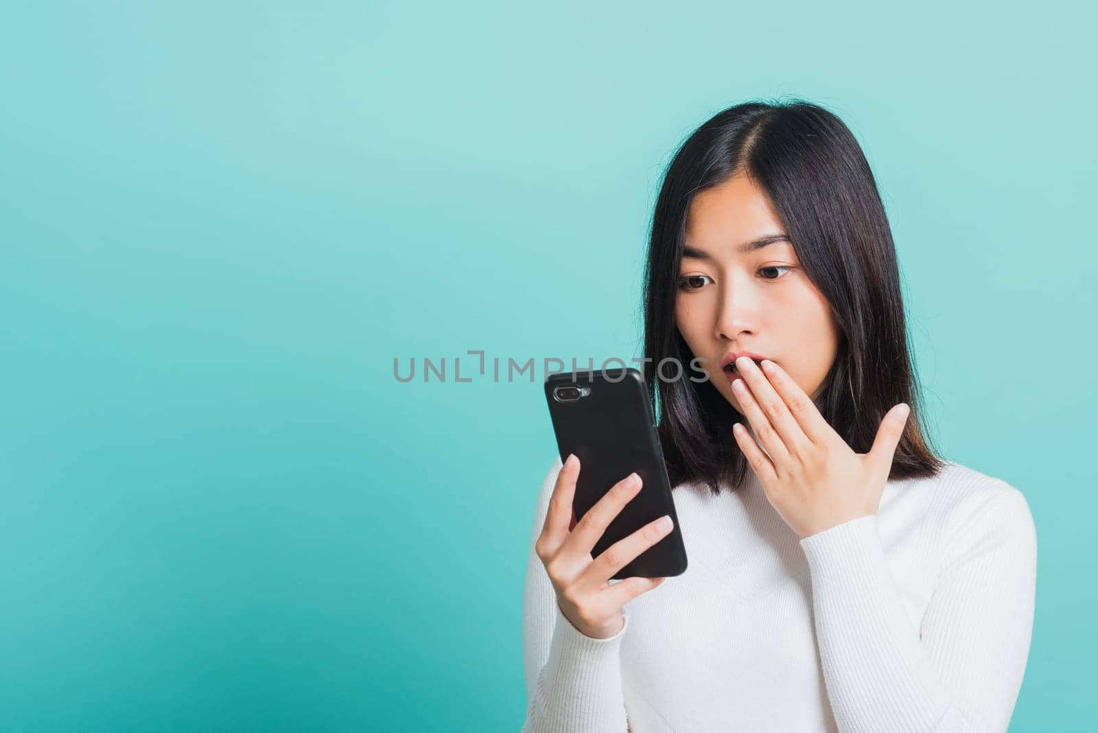 Portrait female anxious scared on the phone seeing bad news, Young beautiful Asian woman surprised shocked with mobile phone close mouth with palm, studio shot isolated on a blue background