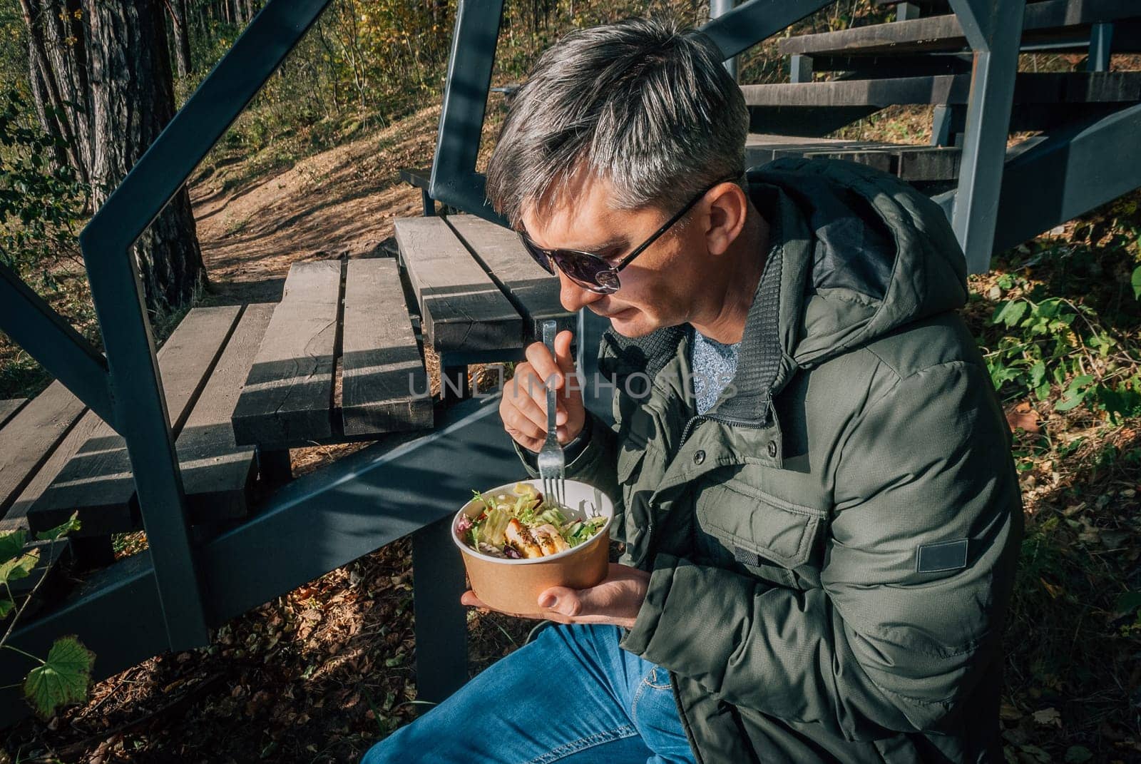 A man dines on a salad from a disposable paper bowl in the open air during a walk in nature in the fall.