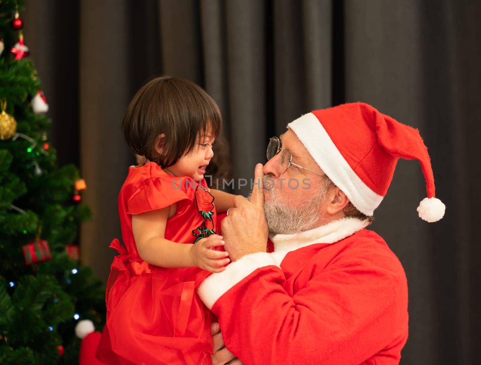 Santa Claus wearing red costume keeping forefinger by his mouth while trying to comfort shouting crying toddler girl near Christmas tree in living room at home