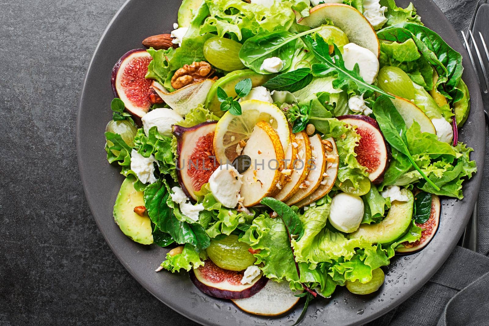 Fresh green leafy salad with mozzarella cheese, fruits and nuts on dark background close up. Concept for a tasty and healthy meal
