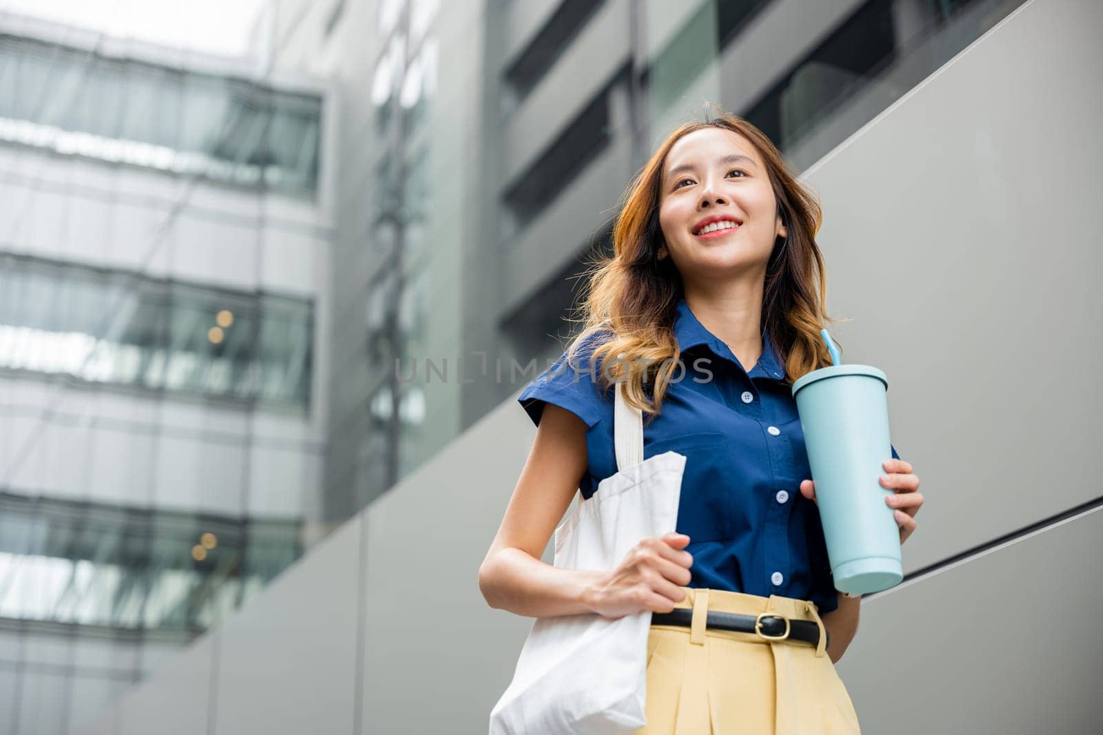 Asian beautiful business woman confident smiling with cloth bag holding steel thermos tumbler mug water glass she walking outdoors on street near modern building office, Happy female