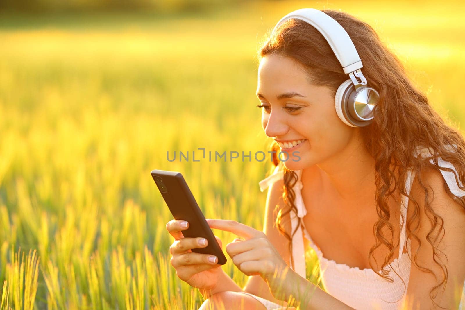 Happy woman in a field listening to music using phone at sunset
