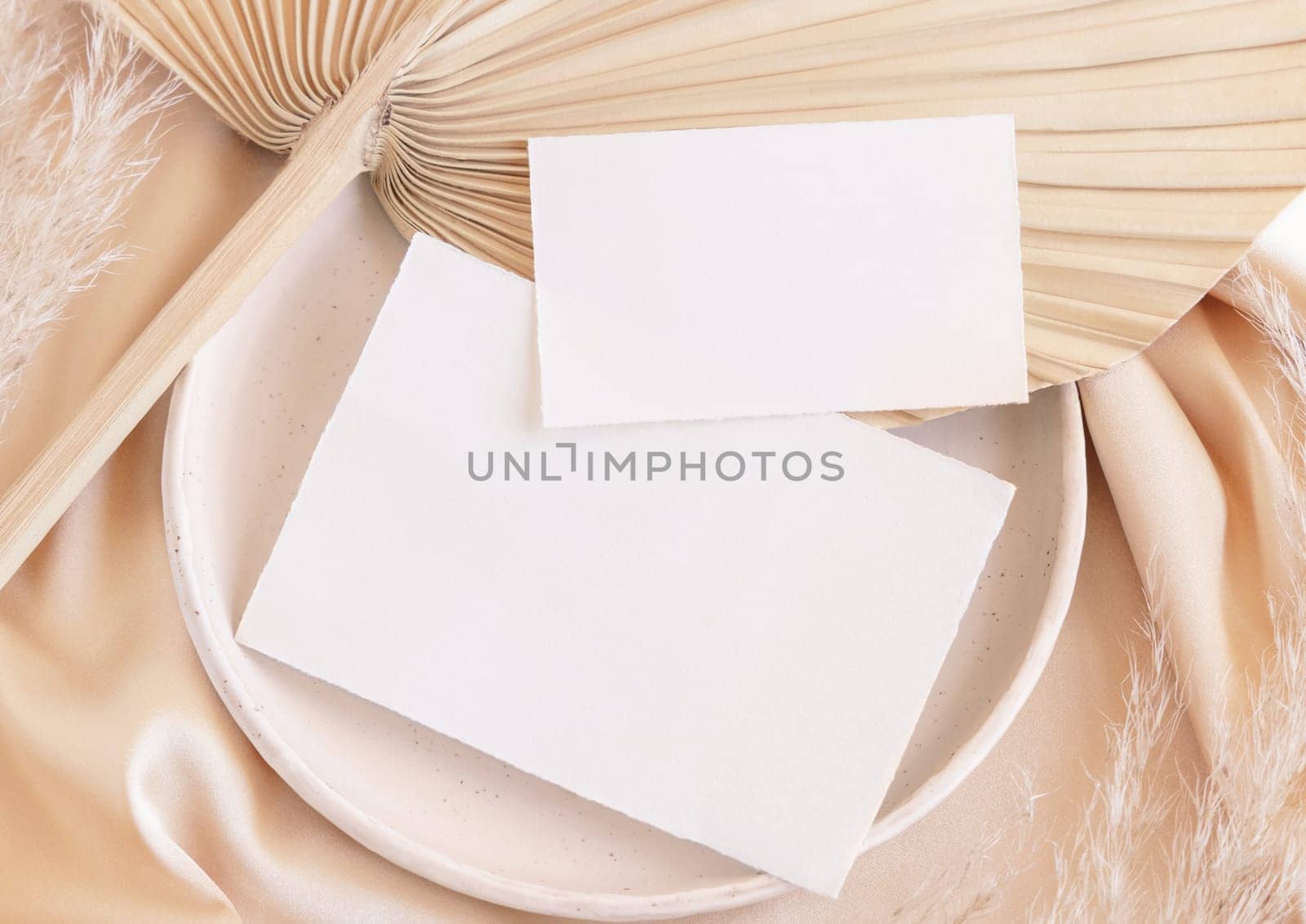 Blank cards on plate with dried palm leaf and beige silky fabric close up, mockup. Romantic scene with horizontal paper card. Bohemian pastel greeting or wedding Invitation .