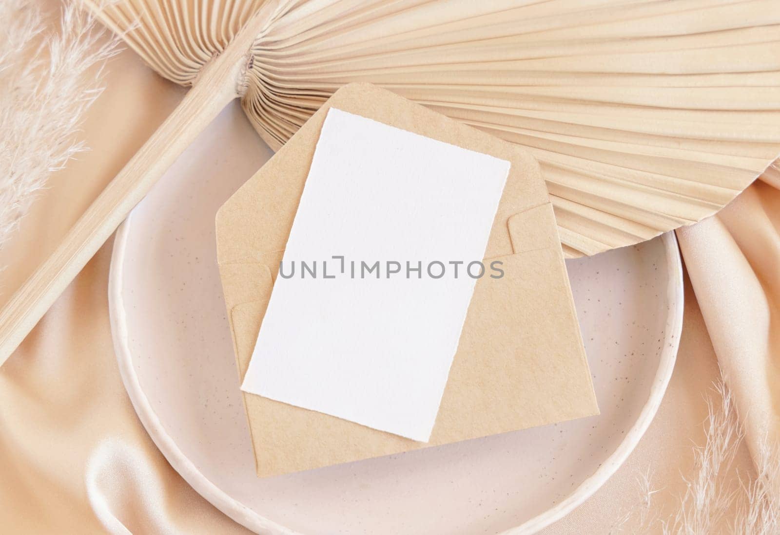 Blank card and envelope on plate with dried palm leaf and beige silky fabric close up, mockup. Romantic scene with vertical paper card. Pastel greeting or wedding stationery.