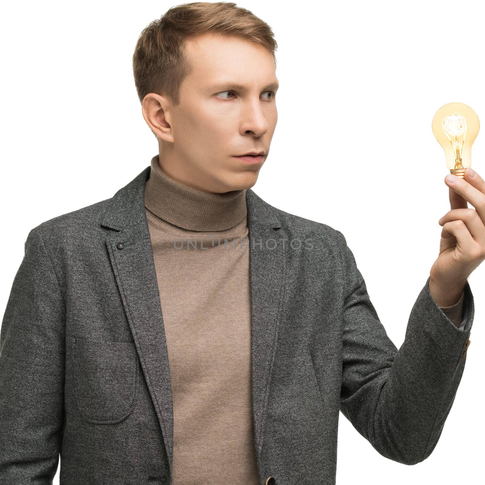 A man dressed in a gray blazer and turtle neck thoughtfully examines a light bulb in a well-lit studio environment.