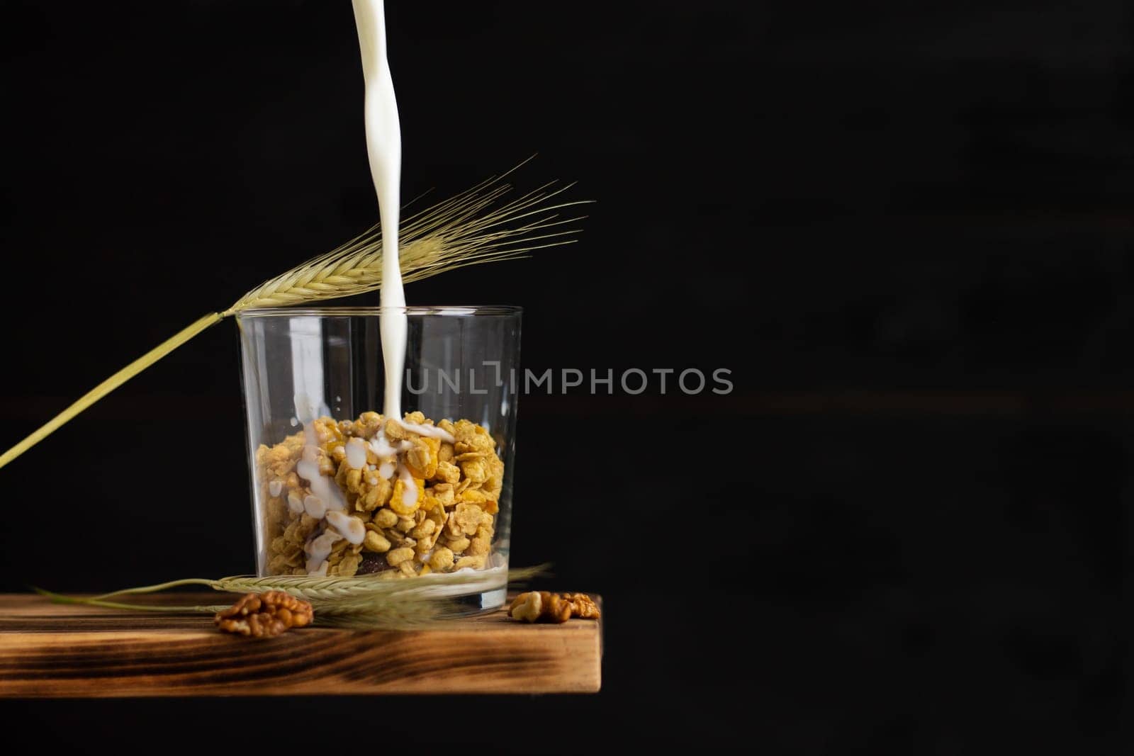 Milk flowing into a cereal granola in a transparent glass with rye spike and walnuts on a shelf on a black background. Copy space.