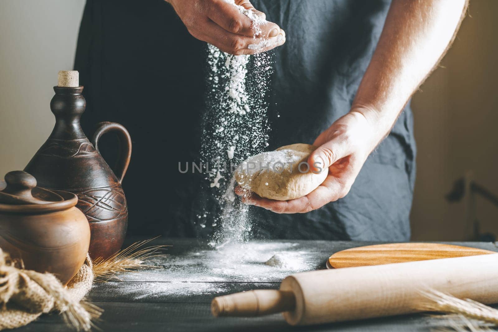 Male hands pour flour on the dough next to clay pot and oil bottle and rolling pin on dark table while cooking