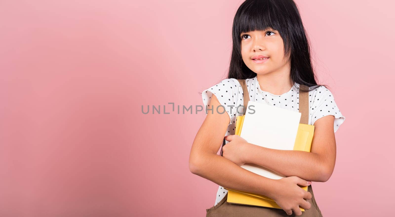 Asian little kid 10 years old holding book for going to school at studio shot isolated on pink background, Happy child girl hugging textbooks dictionary, lifestyle lovely educated cheerful