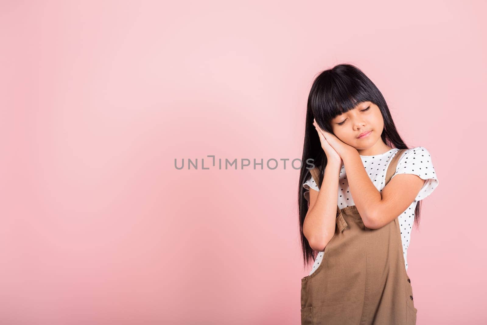 Asian little kid 10 years old standing with hands sealed pretending he is sleeping closed eyes at studio shot isolated on pink background, Happy child girl holding hands under face tired dreaming