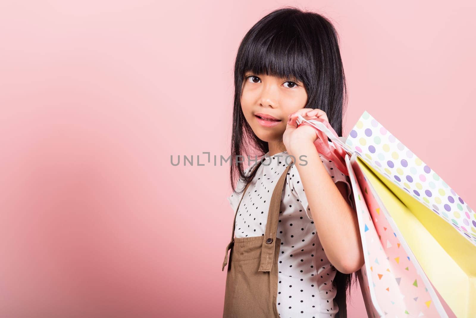 Asian little kid 10 years old smiling holding multicolor shopping bags in hands at studio shot isolated on pink background, Portrait of Happy child girl shopper lifestyle, Black friday concept