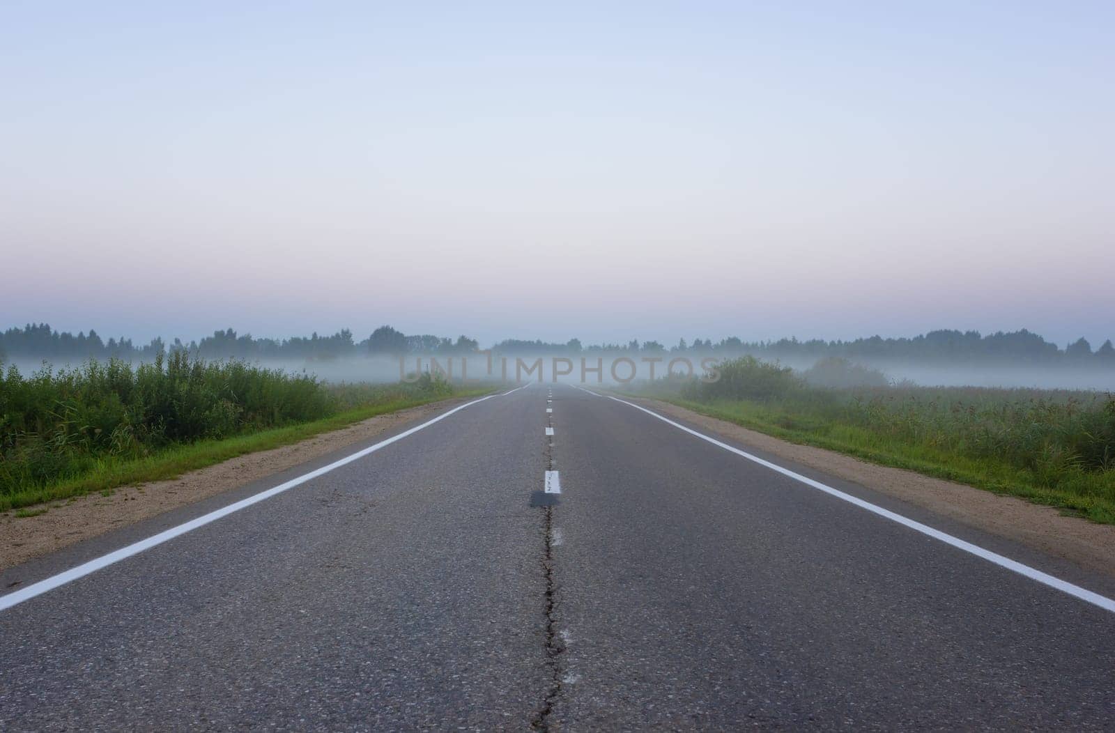 Diminishing perspective view of an empty, straight suburban asphalt roadway with white road markings vanishing into the morning mist at dawn. Yaroslavl Oblast, Russia.