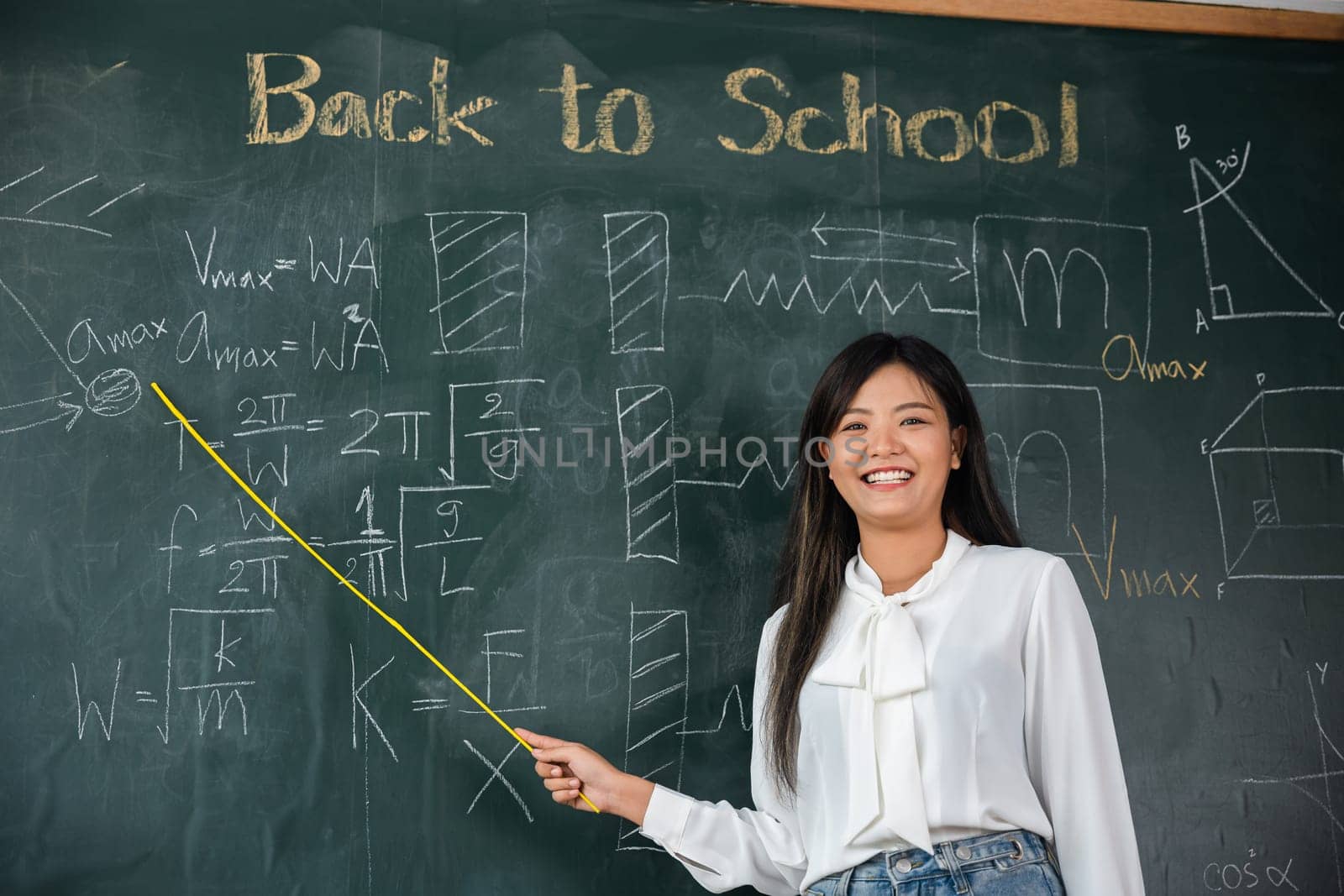 Back to school concept. Happy beautiful young woman standing hold pointer to back board, Asian female teacher smiling with wooden stick pointing to blackboard at school in classroom, Education