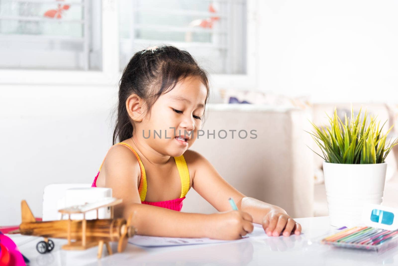 Child little girl drawing cartoon on paper before paint the color, Asian cute kid preschooler sitting on table smiling she draw and writing a picture with pencil at home to learning arts homework