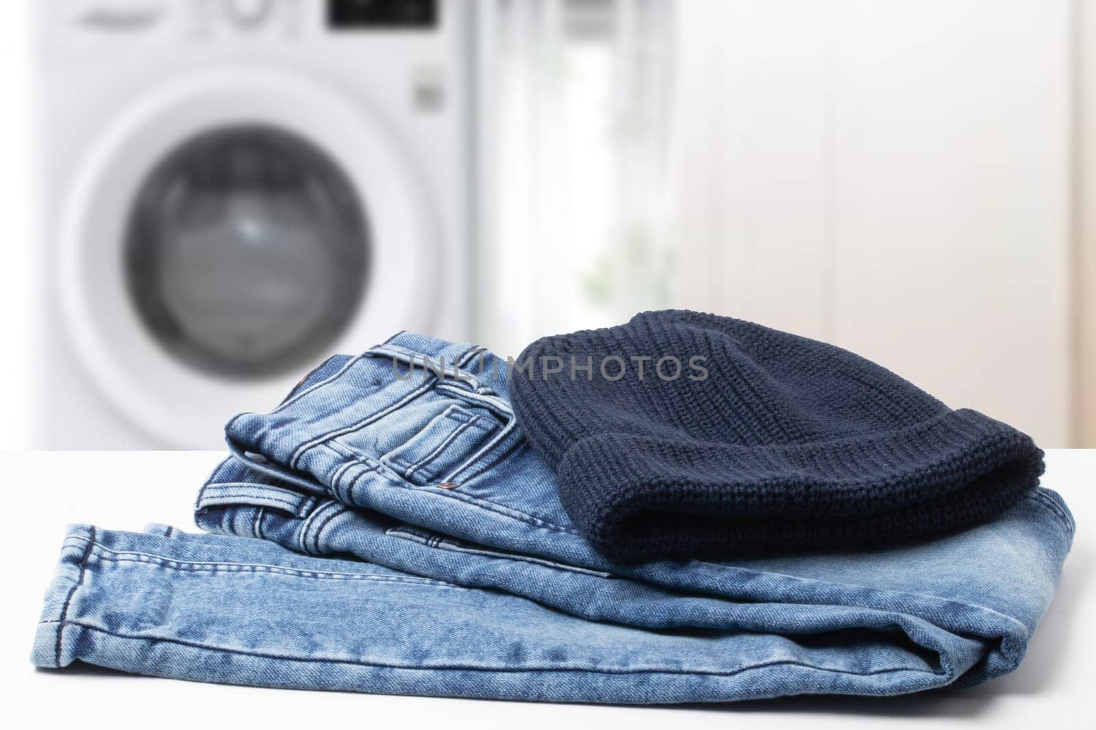 Colorful children jeans clothes. Fashionable folded jeans and a knitted blue Rapper, Beanie or Baseball hat or cap on table over abstract blurred bathroom background.