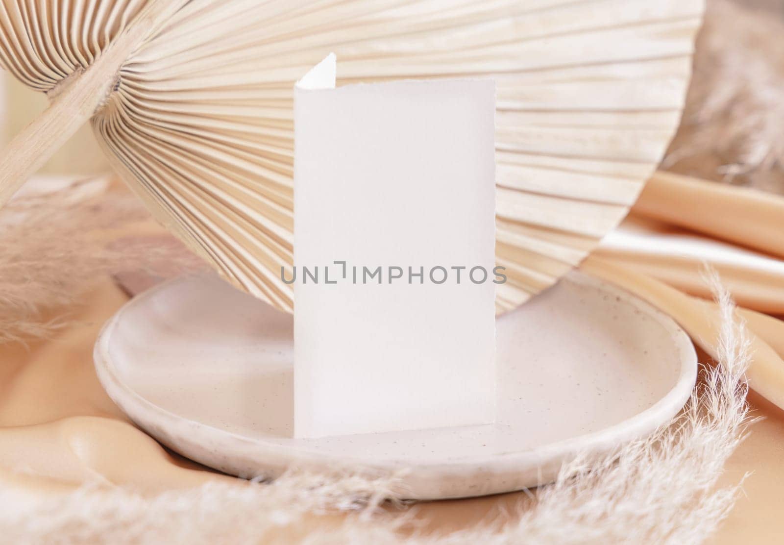 Folded card on plate near dried palm leaf and beige silky fabric close up, table place mockup. Romantic scene with vertical paper card. Pastel greeting or wedding stationery.