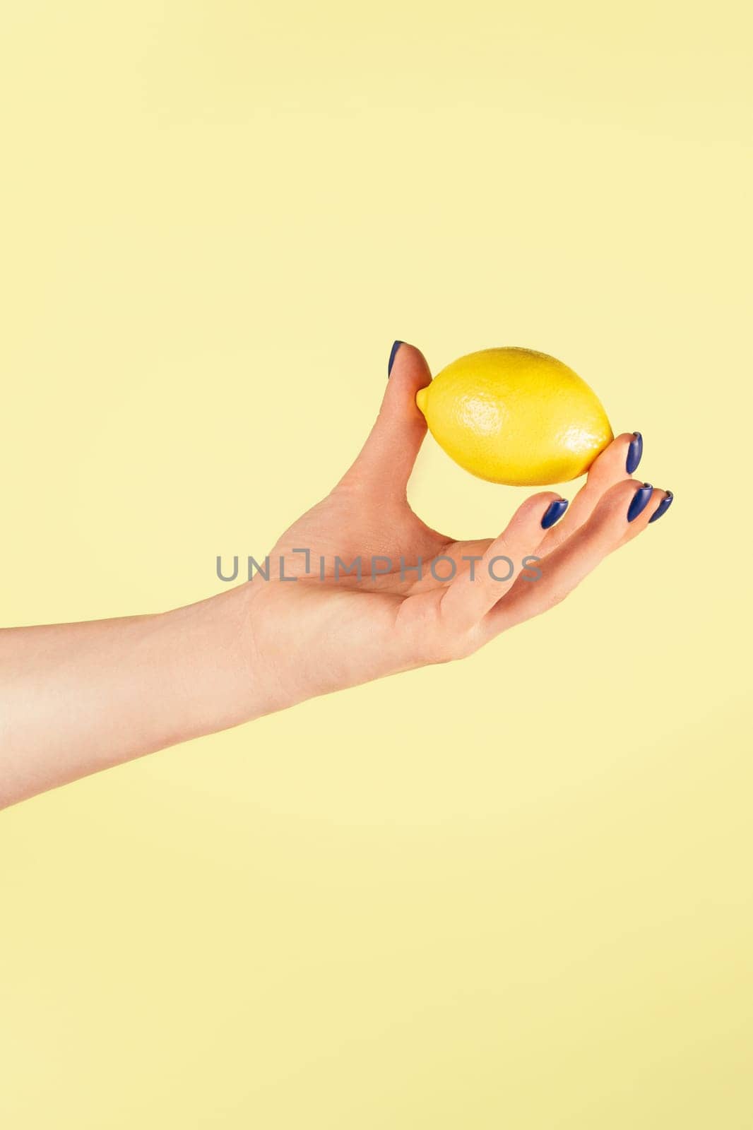 Female hand with blue nails holding slightly with two fingers fresh ripe juicy lemon isolated on yellow. Creative art concept. Food minimal colorful concept. Vertical. Copy space. Mock up.