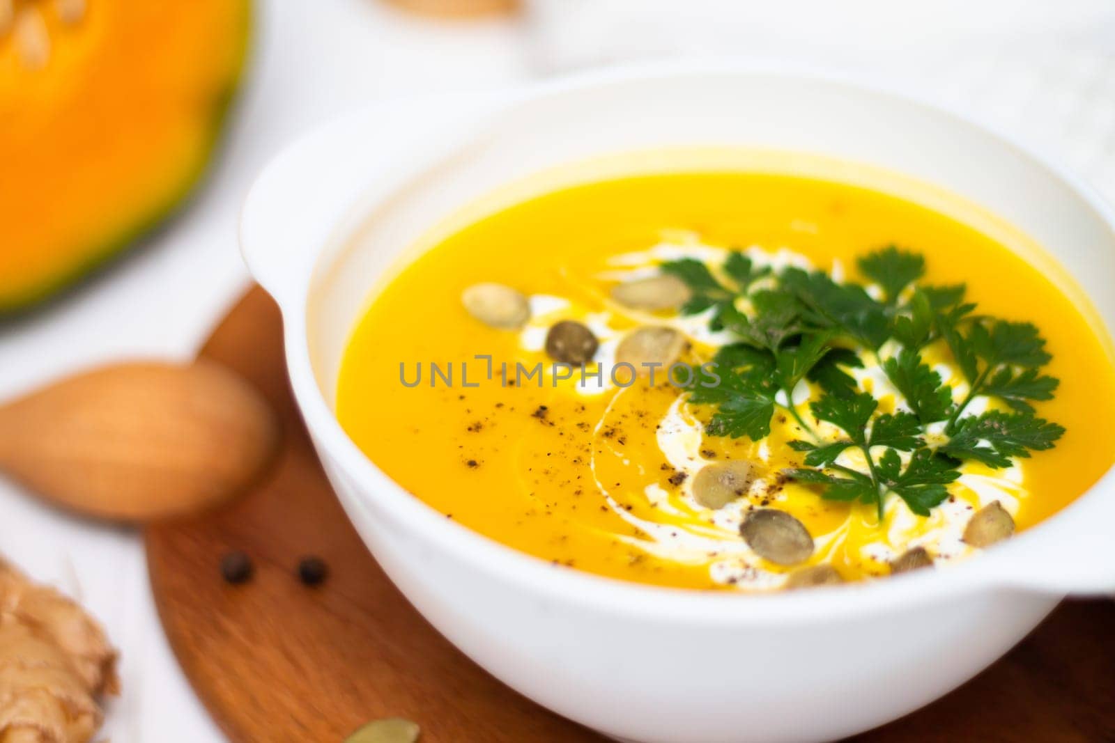 A plate of yellow pumpkin cream soup with cream, herbs and seeds on a white table near ginger, raw pumpkin and a wooden spoon.