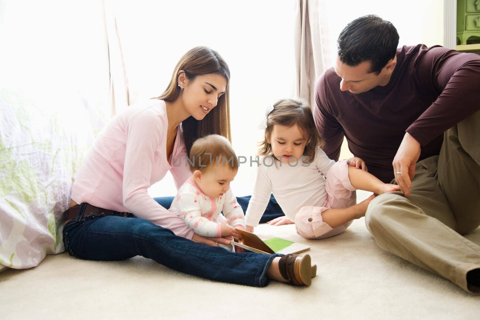 Caucasian girl children with mother and father sitting on bedroom floor looking at book.