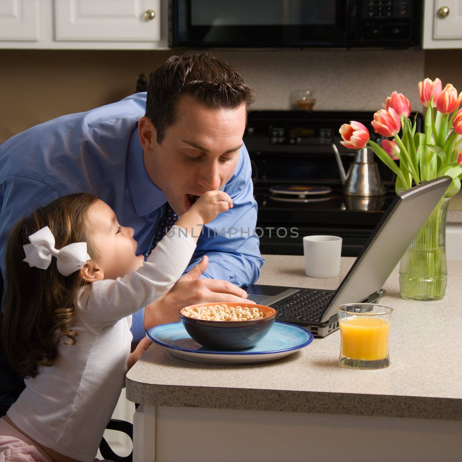 Caucasian father in suit using laptop computer with daughter feeding him breakfast in kitchen.