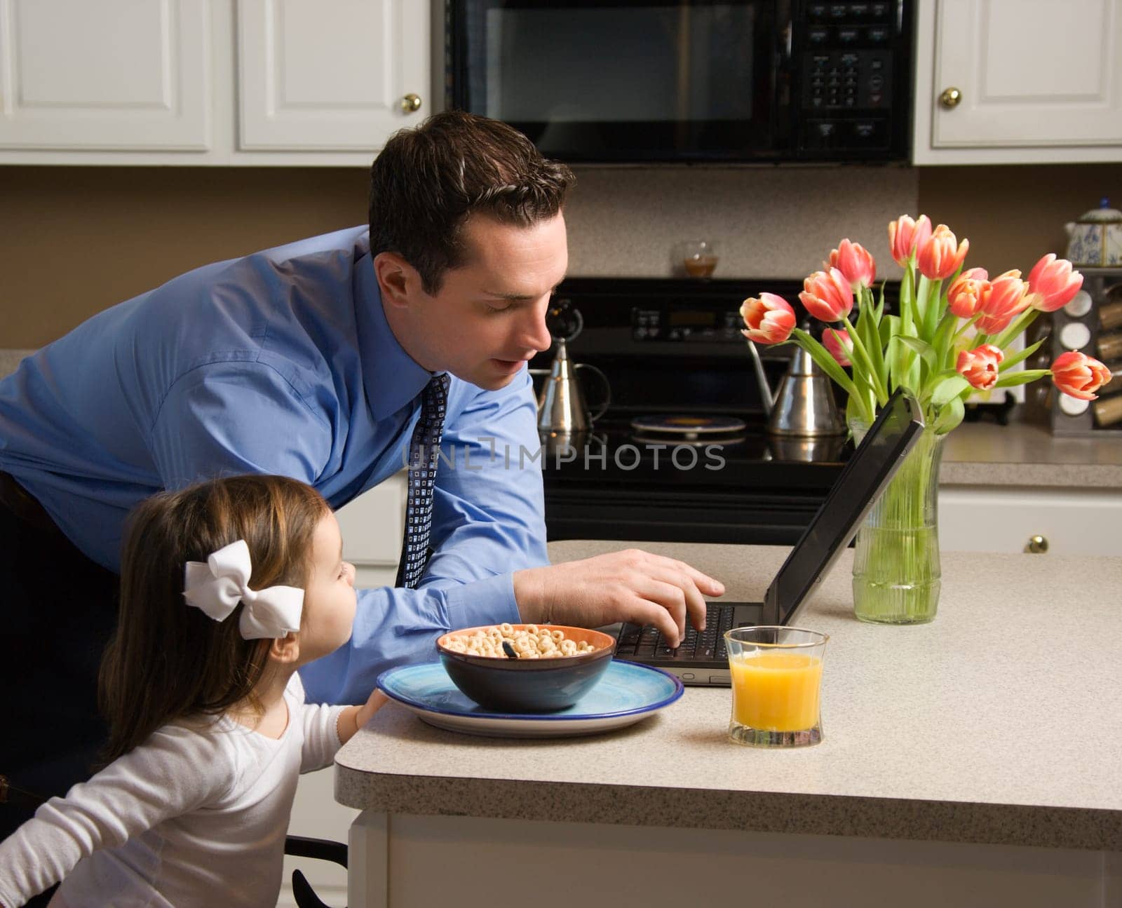 Caucasian father in suit using laptop computer with daughter eating breakfast in kitchen.