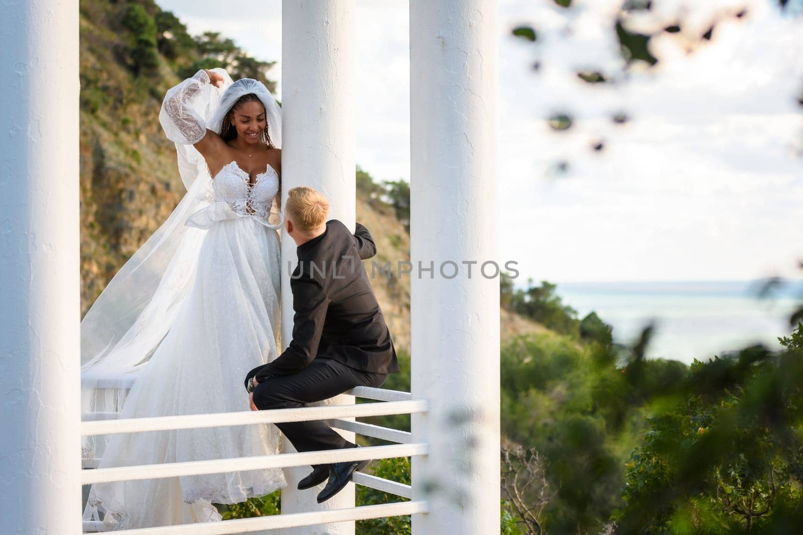 The newlyweds are walking in a beautiful picturesque gazebo, the girl is holding a veil with her hand, the guy is sitting on the railing