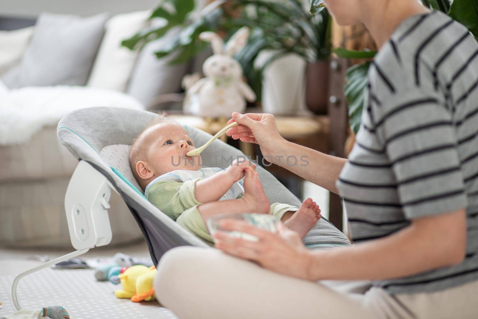 Mother spoon feeding her baby boy child in baby chair with fruit puree. Baby solid food introduction concept