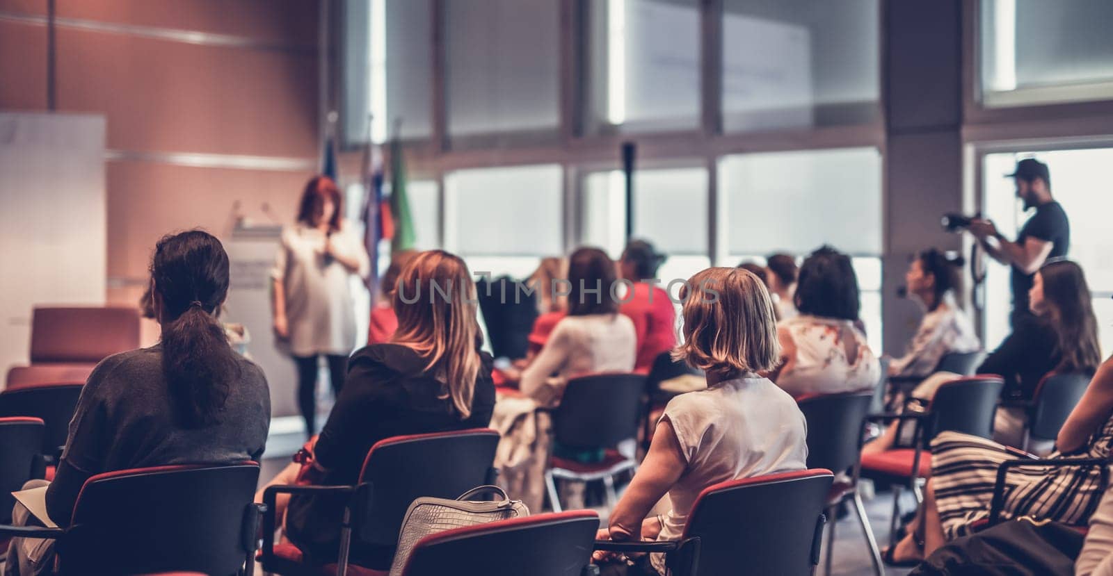 Business and entrepreneurship symposium. Female speaker giving a talk at business meeting. Audience in conference hall. Rear view of unrecognized participant in audience.